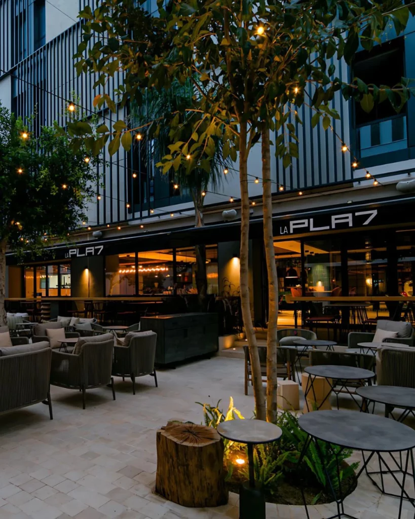 Vue extérieure de la terrasse La Pla7 avec tables, chaises, arbres et guirlandes lumineuses en soirée